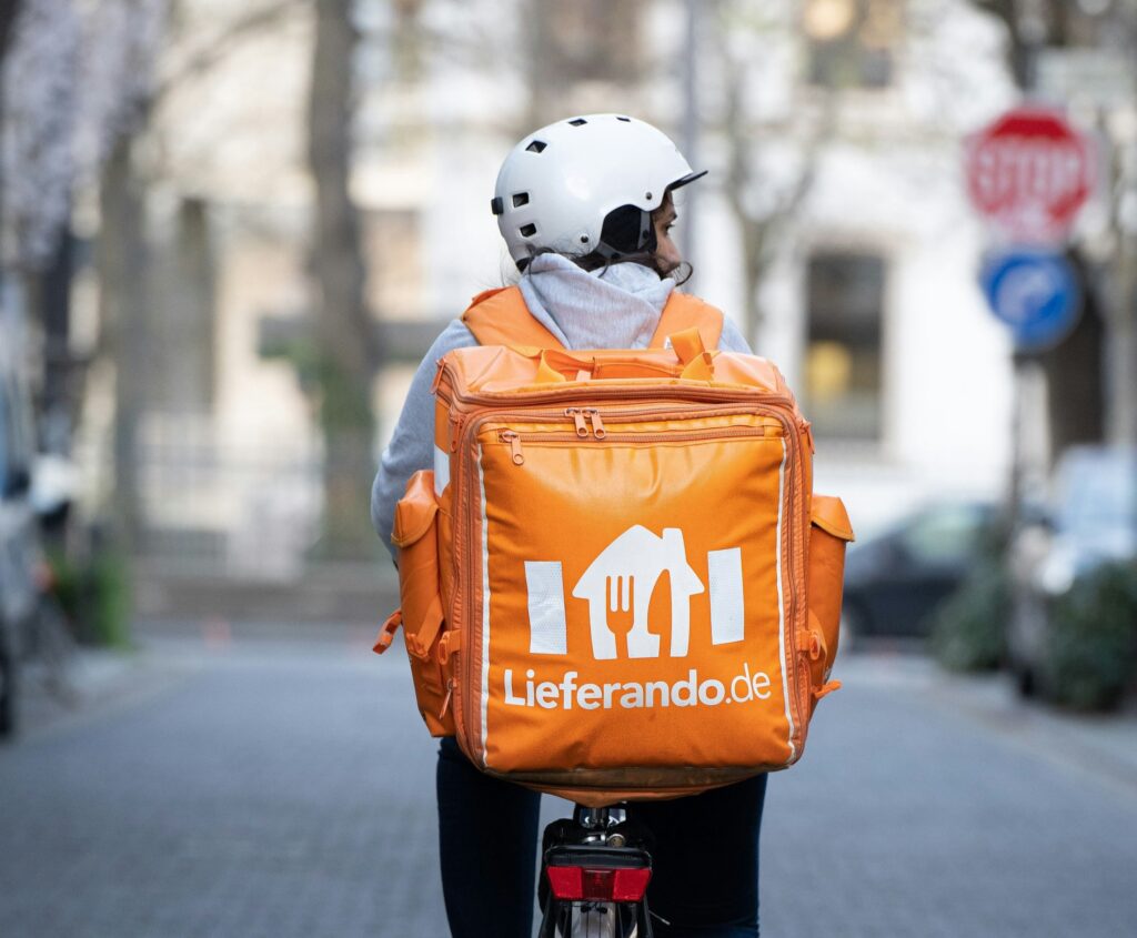 Lieferando-Fahrer:in auf Fahrrad mit orangefarbener Lieferbox fährt durch eine städtische Straße.
